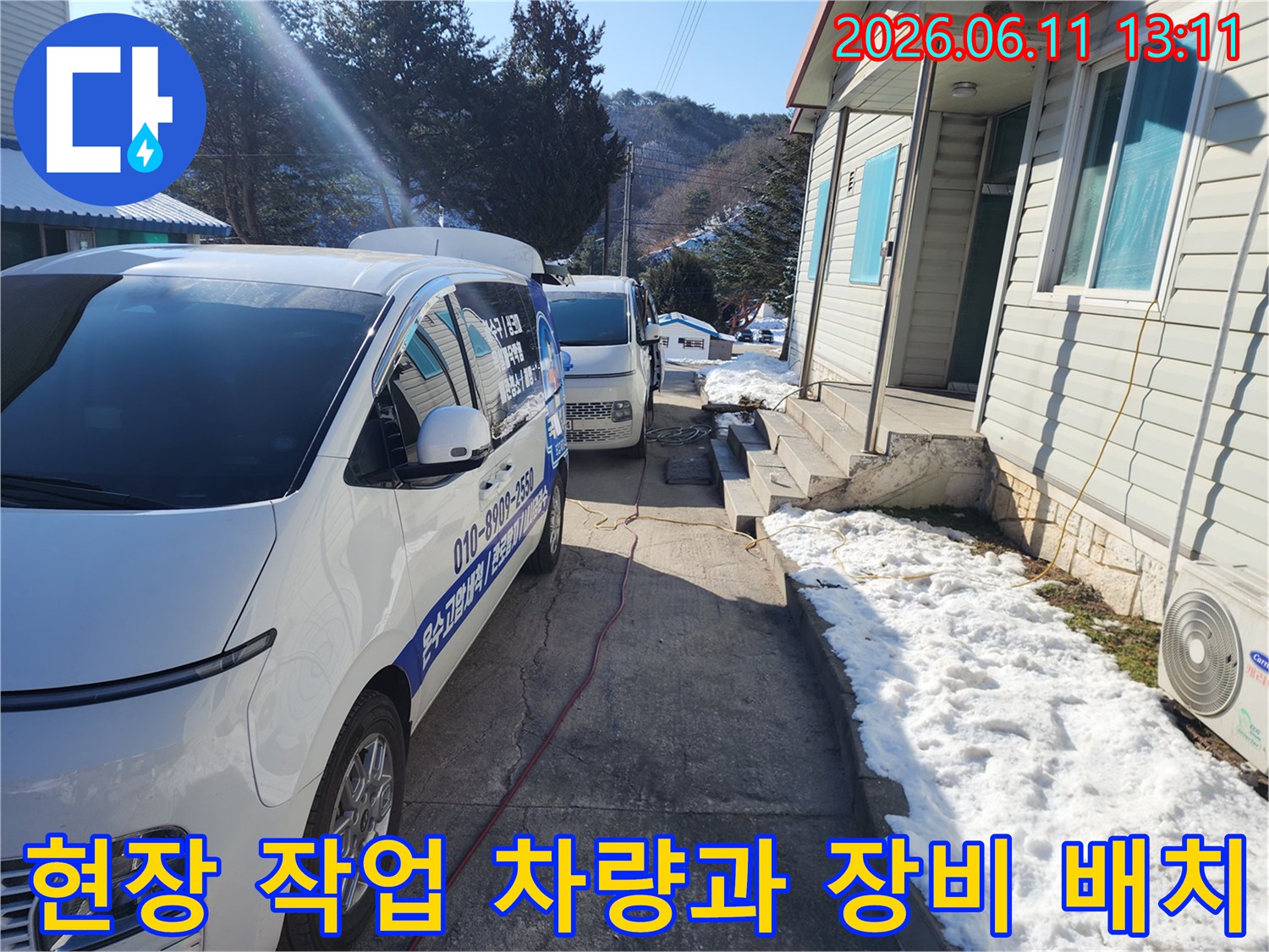 눈 덮인 주택가 건물 옆에 주차된 작업용 차량과 배관 연결 호스입니다.