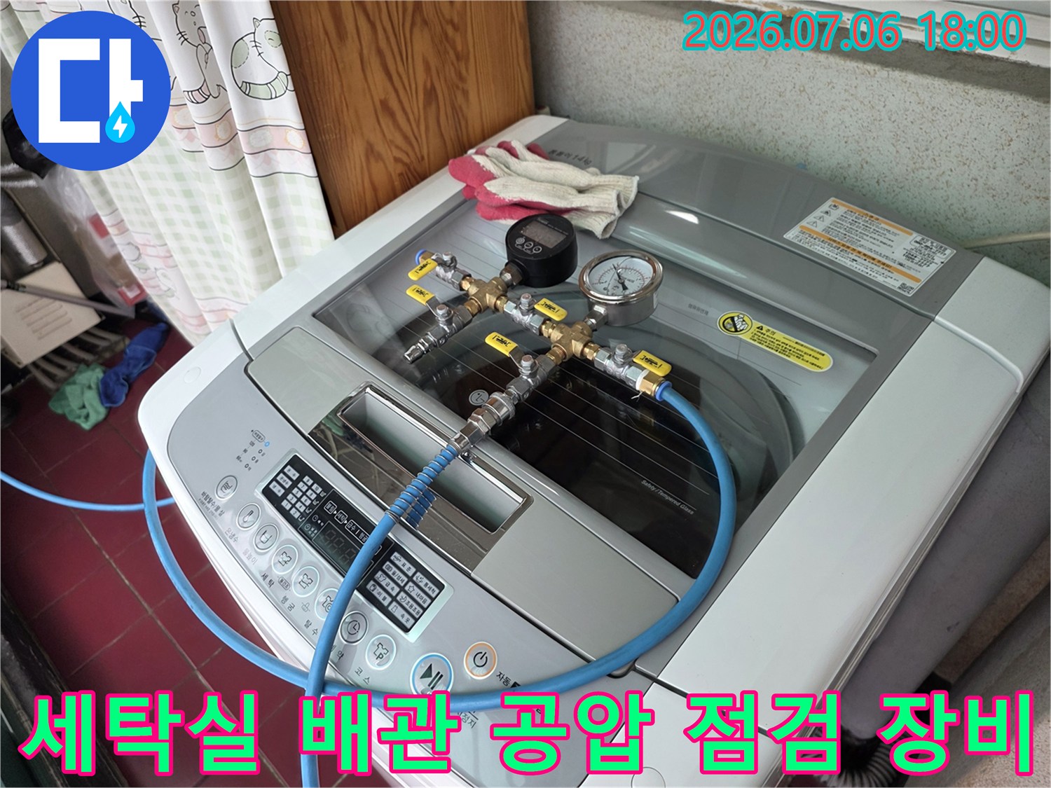 세탁실 내부에 설치된 세탁기 위로 연결된 파란색 호스와 게이지가 달린 압력 측정 장치