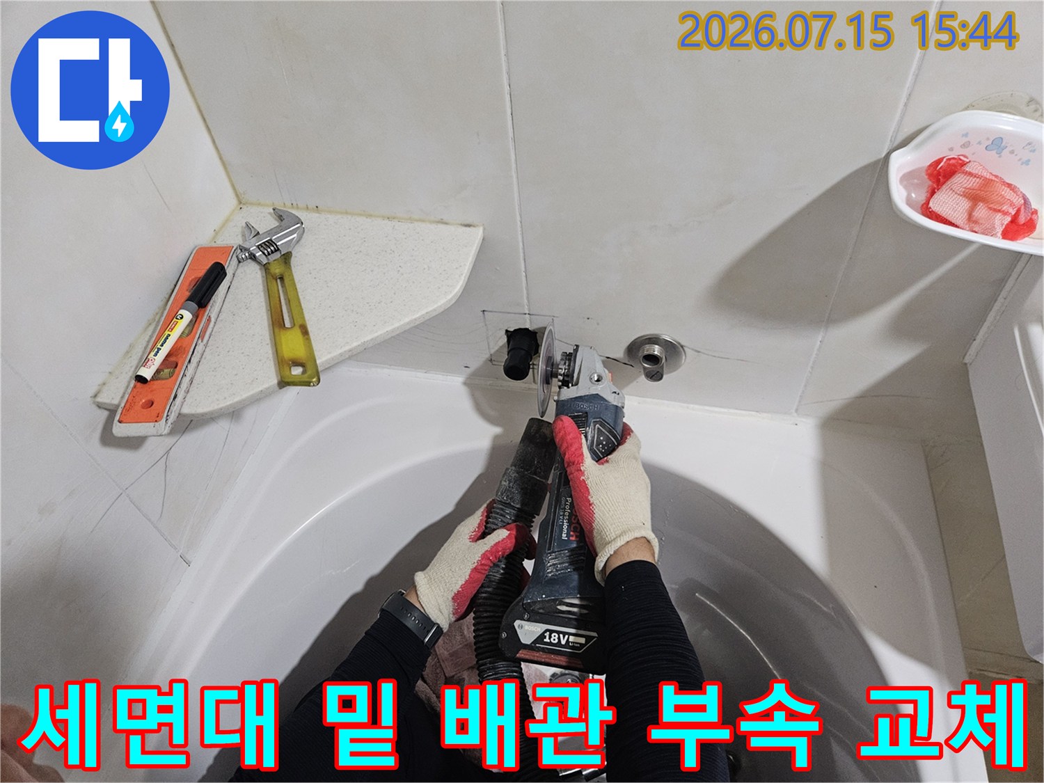 작업자가 장갑을 끼고 세면대 아래 노출된 수도 배관 연결부를 수리하는 사진